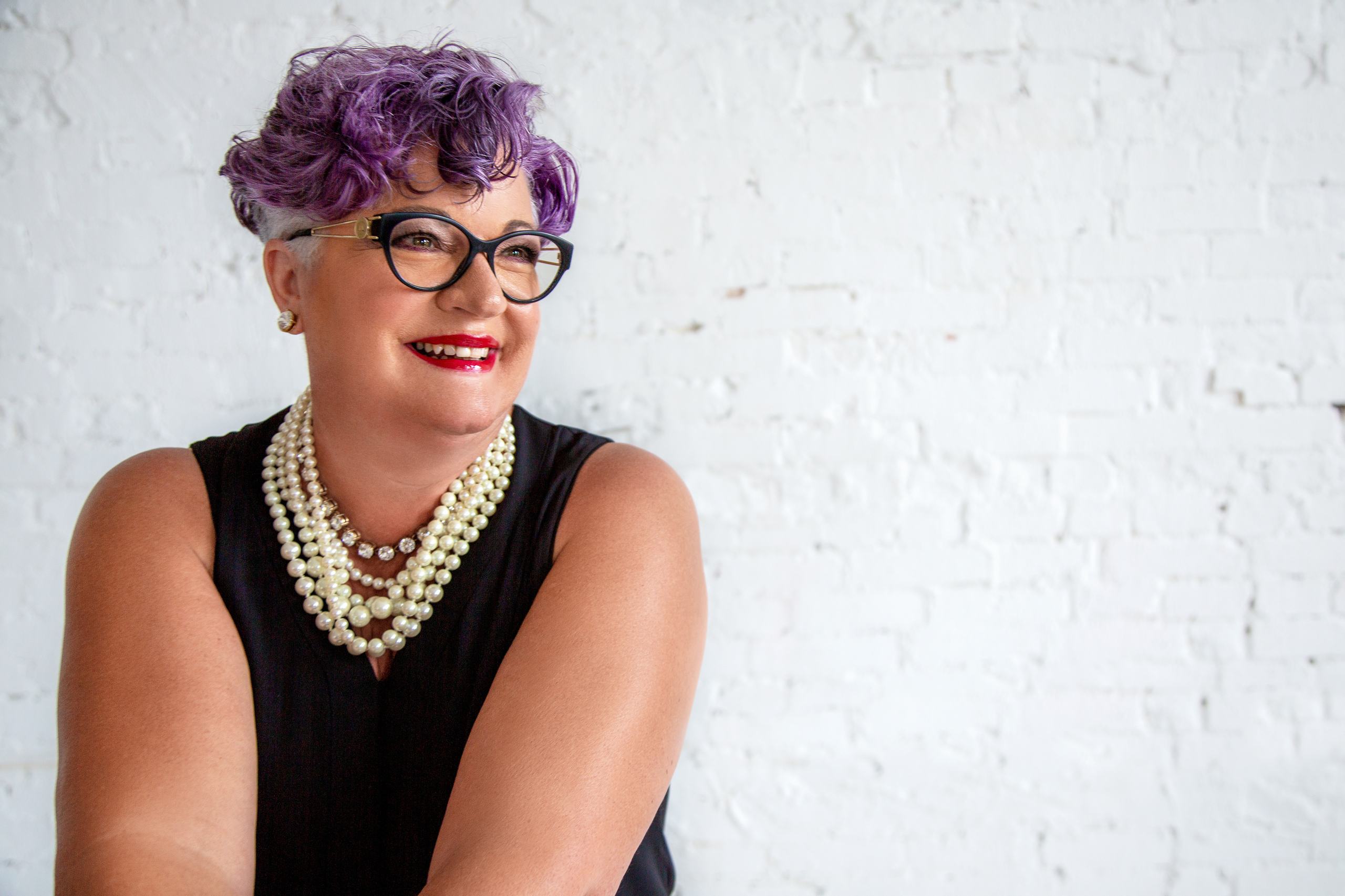 Brenda Florida, in a black top and layered pearls, smiling in front of a white painted brick wall